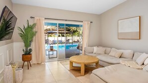 Una sala de estar moderna con un sofá beige, una mesa de centro redonda de madera y un ventanal con vista a una piscina.