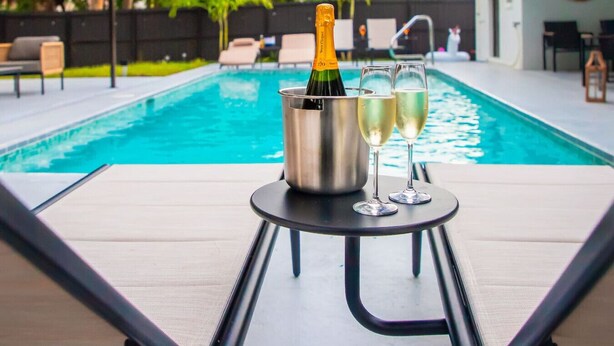 Escena junto a la piscina con una botella de champán y dos copas sobre una mesita.