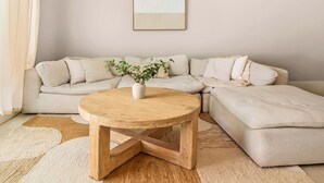 Una sala de estar con un sofá seccional color beige, una mesa de centro redonda de madera con una planta, y una pared lisa con un cuadro enmarcado.