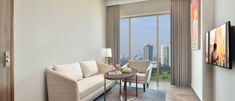 A modern hotel room with a sofa, armchair, and a table with a drink. Large windows offer a city view.