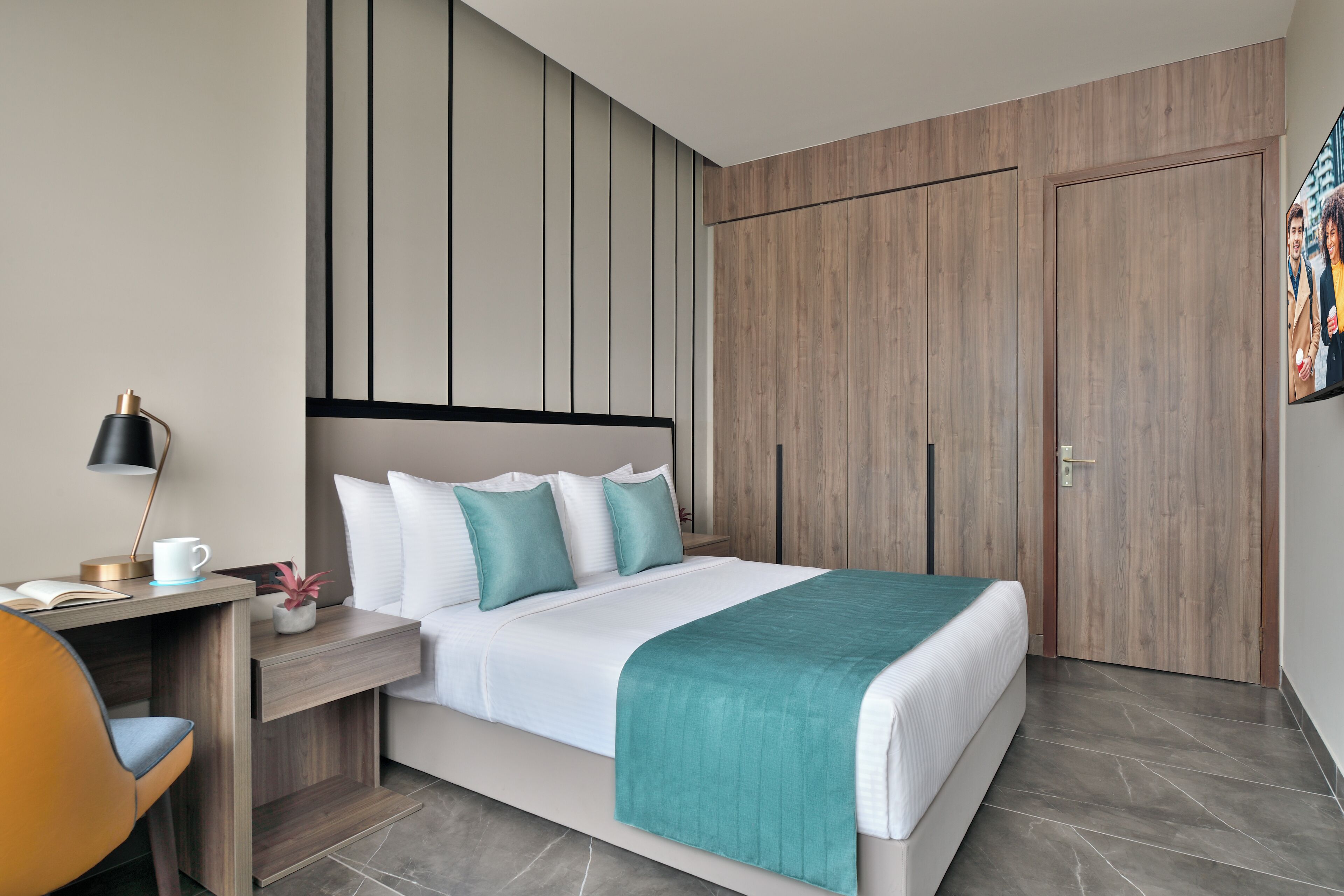 A modern hotel room with a bed, a desk with a lamp, a chair, and a wardrobe.