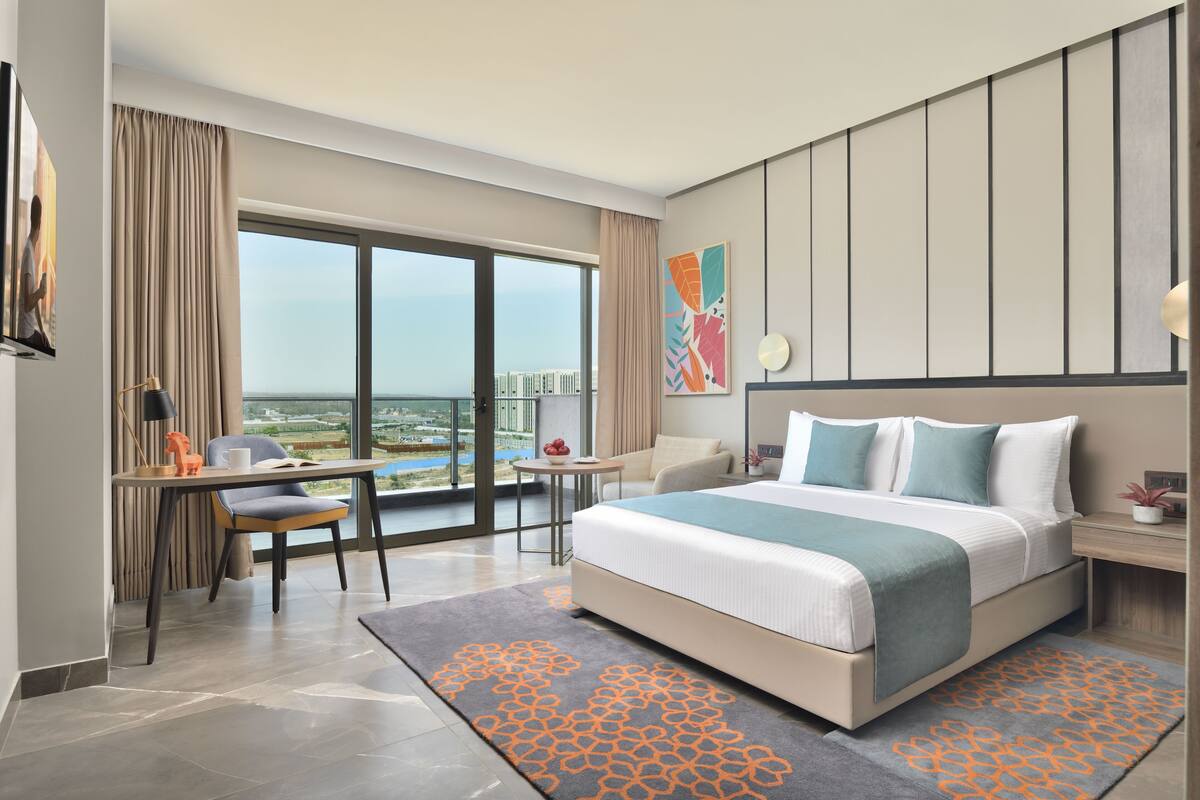 Ein modernes Hotelzimmer mit einem großen Bett, einem Schreibtisch mit Stuhl, einem kleinen Tisch und Blick auf die Stadt durch große Fenster.