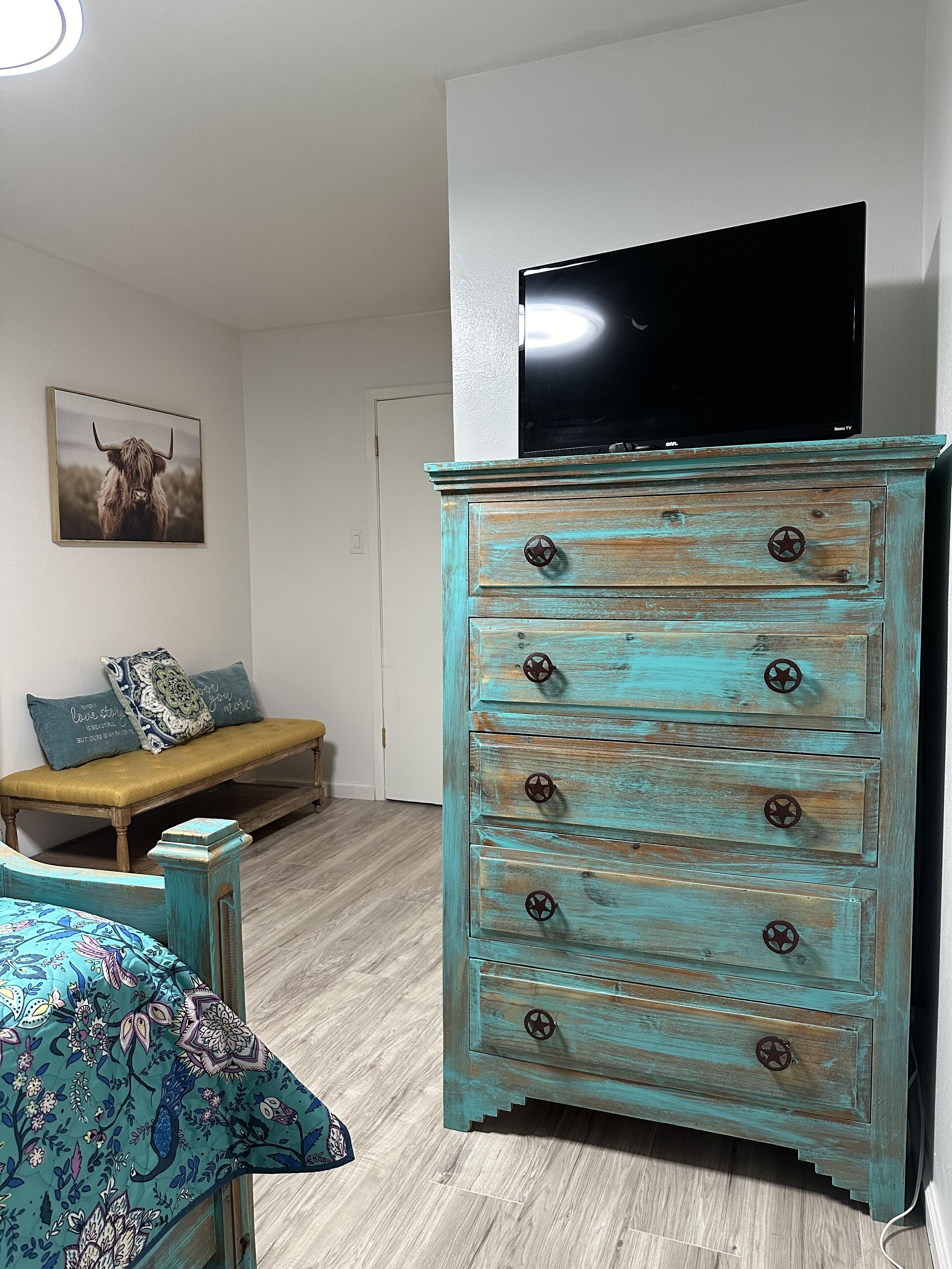 A teal dresser with drawers and a flat-screen TV on top, a bench with cushions, and a framed picture on the wall.