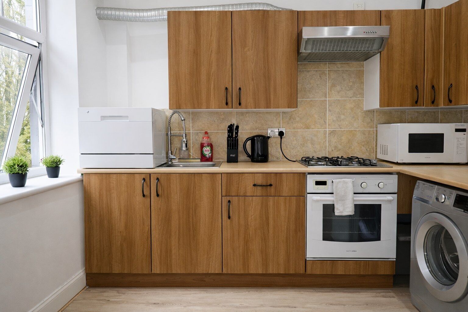 A modern kitchen with wooden cabinets, a stainless steel range hood, a microwave, a gas stove, a washing machine, and a sink with a kettle.