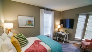A hotel room with a bed, a desk, a chair, a TV, and a window with blinds. - The Capitol Inn - Destin Room - Queen bed (Tallahassee)