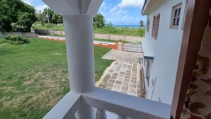 Vista desde un balcón con baranda blanca, sobre un jardín césped y un edificio con puerta roja.