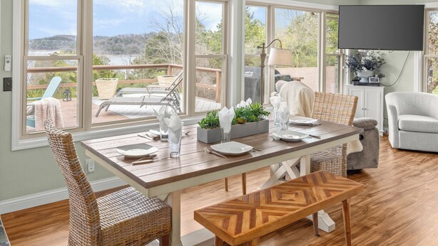 A dining area with a table set for four, large windows, wicker chairs, a bench, and a view of a deck and trees.