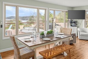 A dining area with a table set for four, large windows, wicker chairs, a bench, and a view of a deck and trees. - Rambo's Adventure & Relaxation (Rogers)