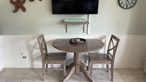 A dining area with a round wooden table, two chairs, a flat-screen TV mounted above, a wall clock, and two decorative starfish on the wall.