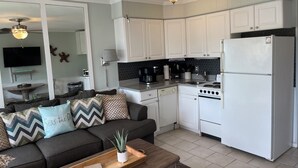 A compact living space with a kitchenette, a sofa with patterned cushions, a ceiling fan, and a flat-screen TV mounted on the wall.