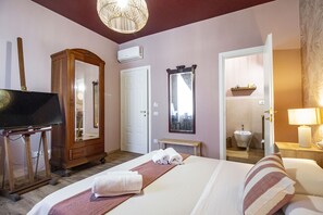 Habitación de hotel con una cama, un armario de madera, un televisor sobre un pedestal, un espejo, un banco y un baño visible a través de una puerta abierta.