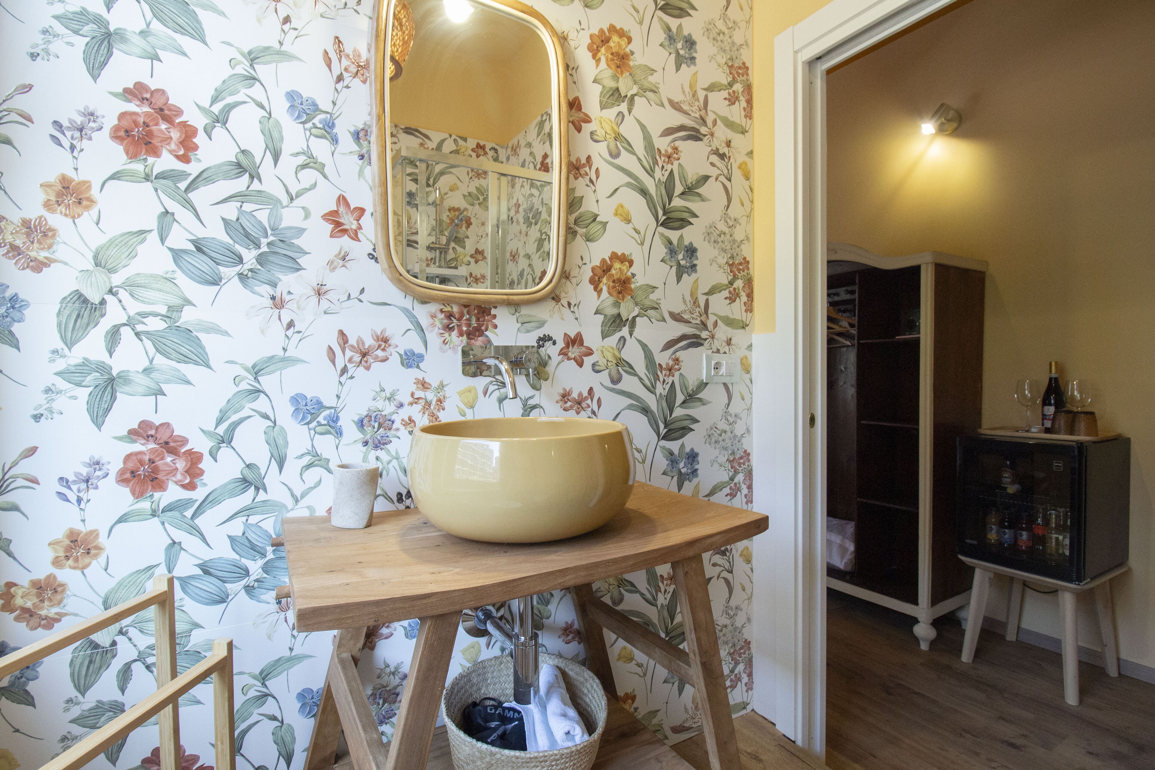 Un baño con un lavamanos grande tipo recipiente, tocador de madera, papel tapiz floral y un espejo.