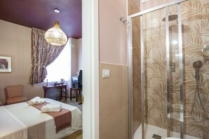 Habitación de hotel con cama, escritorio, silla, ducha con puerta de vidrio y lámpara colgante.