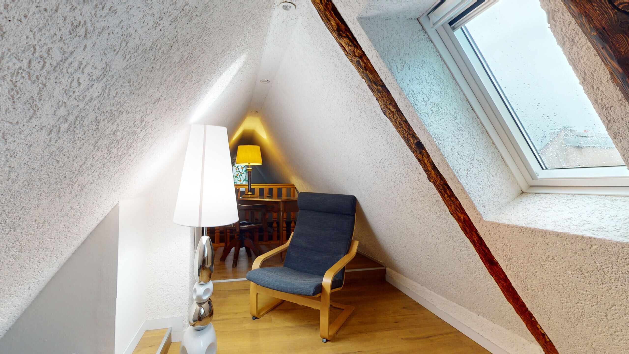 Un coin de pièce avec un plancher en bois, une chaise bleue, une lampe et une fenêtre.