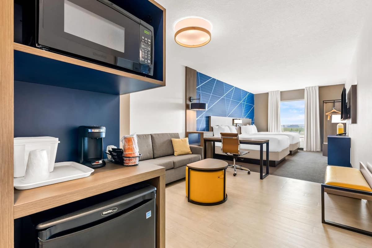 A modern hotel room with a kitchenette, a sofa, a dining table, a small round table, a bed, and a view of the outdoors.