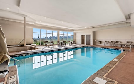 An indoor swimming pool with clear blue water, surrounded by a tiled deck, and a seating area with chairs and tables.