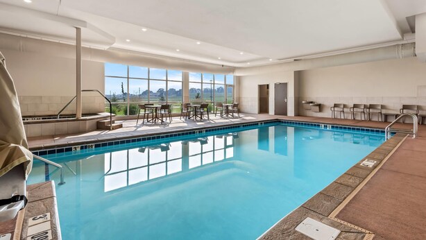 An indoor swimming pool with clear blue water, surrounded by a tiled deck, and a seating area with chairs and tables. - Comfort Inn & Suites at Sanford Sports Complex (Sioux Falls)