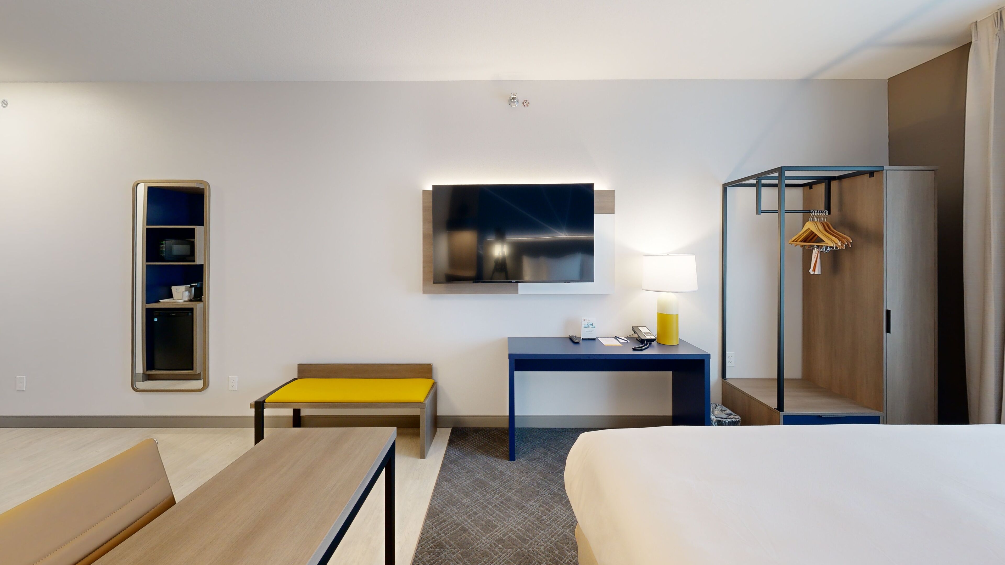 A hotel room with a bed, a desk with a lamp, a TV, a small table, a bench, a wardrobe, and a shelf with items.