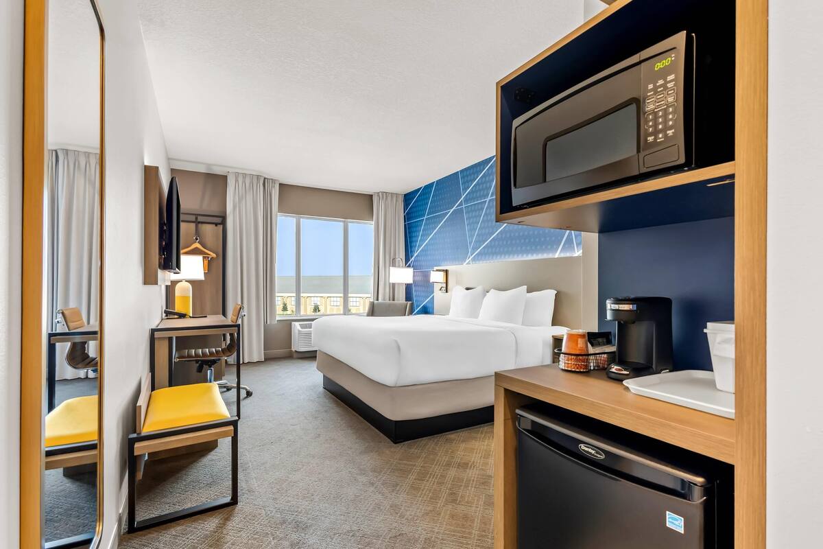 A hotel room with a bed, a desk with a chair, a microwave, a coffee maker, and a mini-fridge.