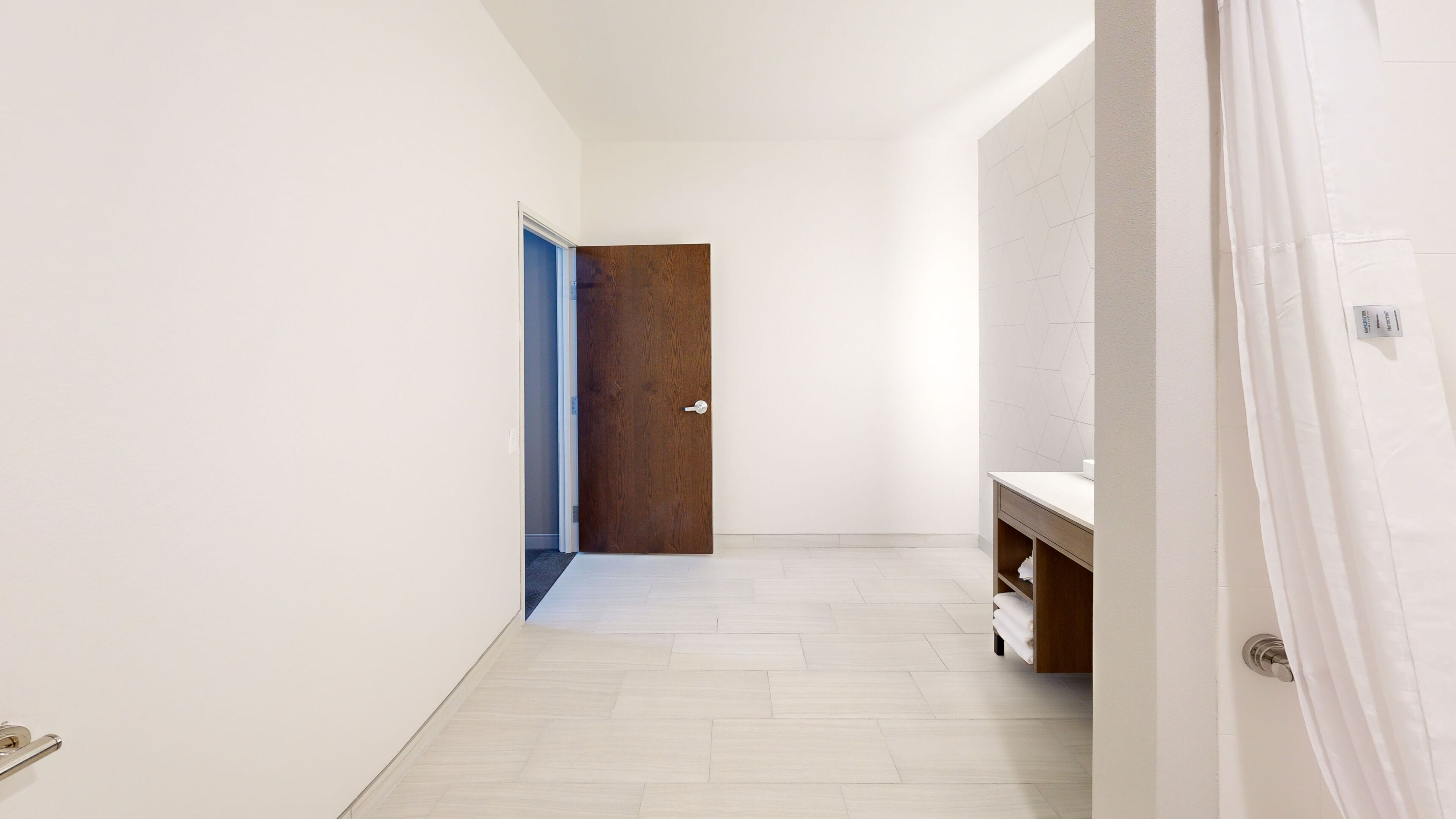 A hallway with a wooden door, a white tiled floor, and a white wall.