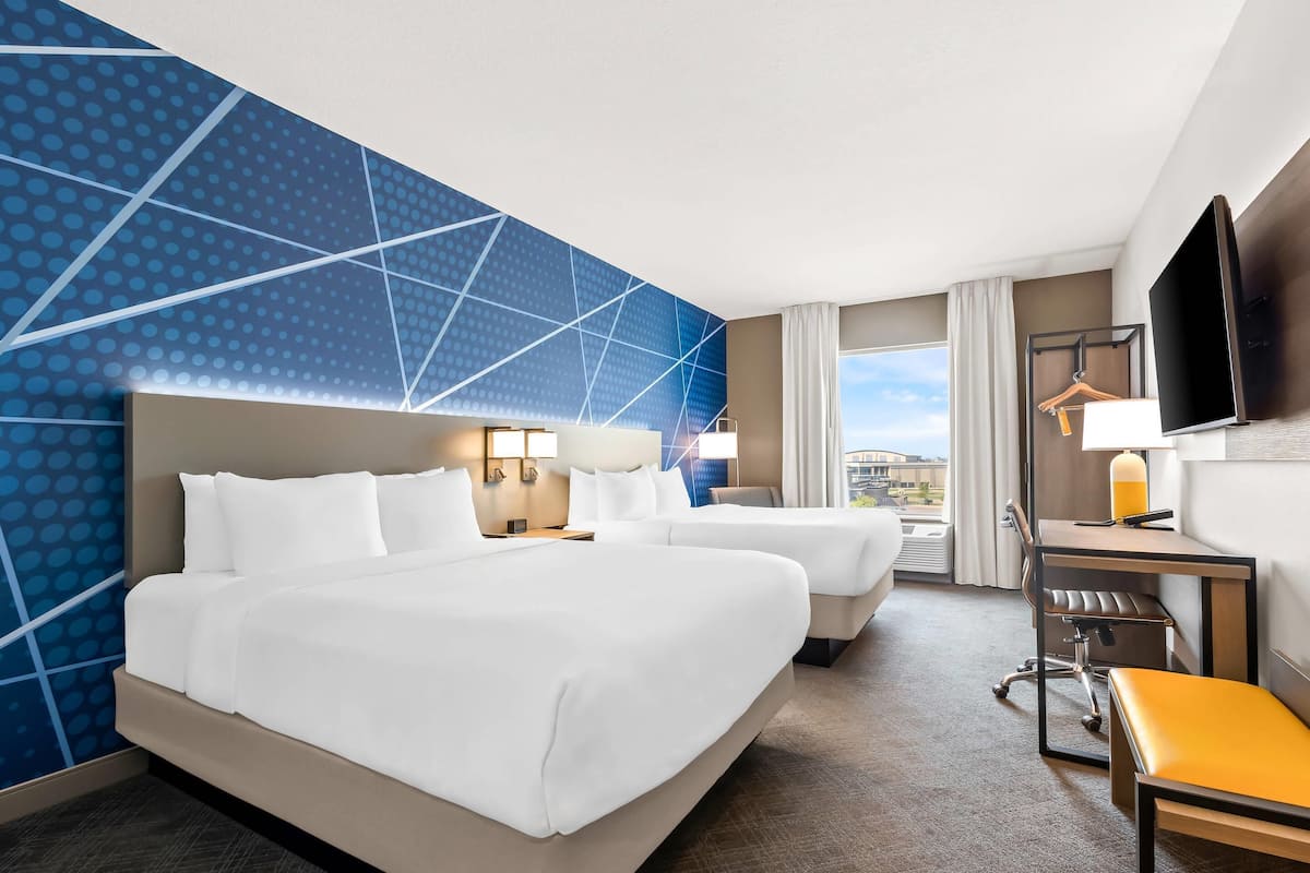 A hotel room with a large bed, a desk, and a chair. There is a TV mounted on the wall and a window with a view of the city.