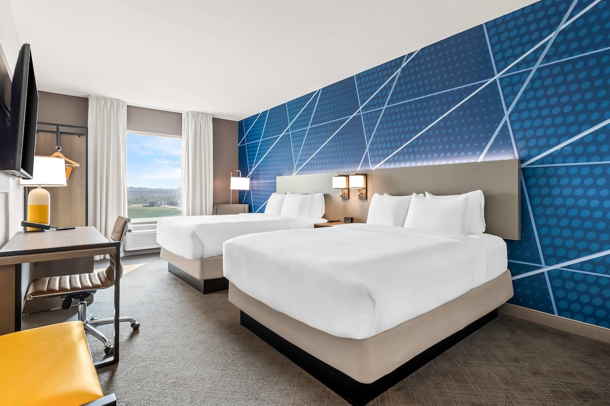 A hotel room with two beds, a desk, a chair, a TV, and a large window with a view.