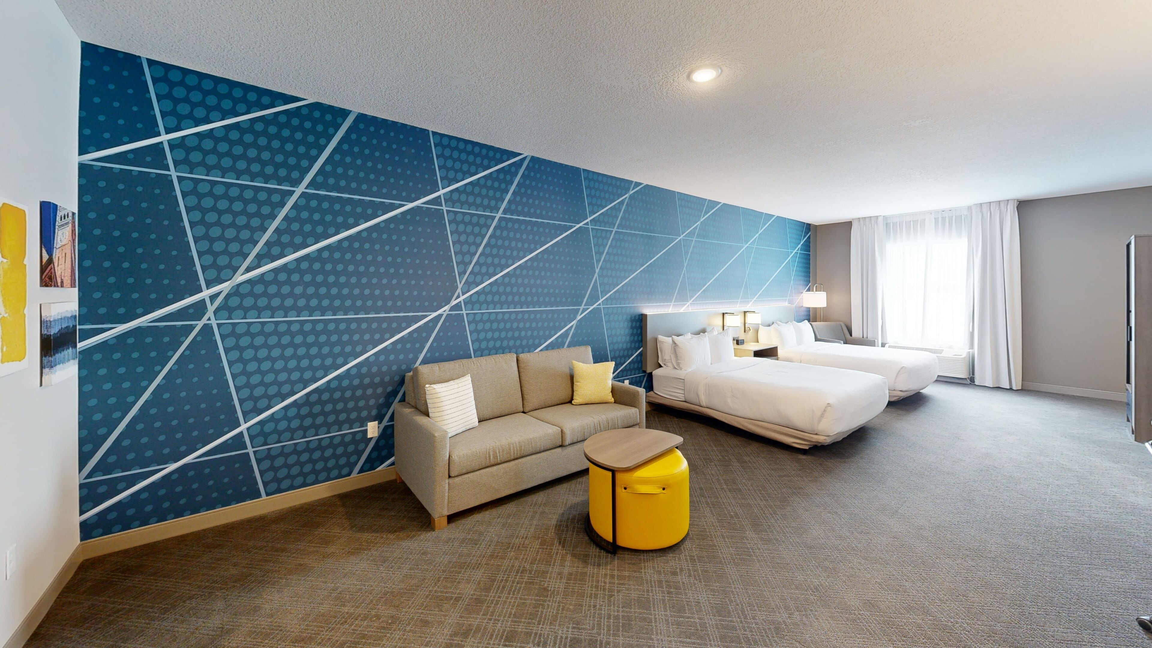 A hotel room with a geometric patterned wall, a sofa, a bed, a bedside table, and a yellow side table.