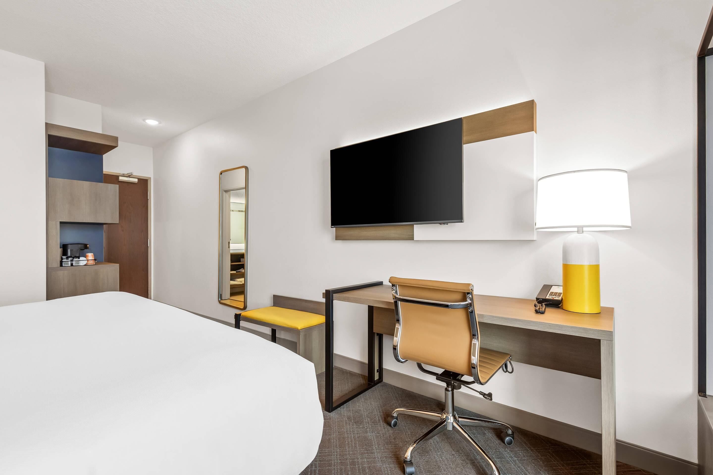 A hotel room with a bed, a desk with a chair, a television, a mirror, and a lamp.