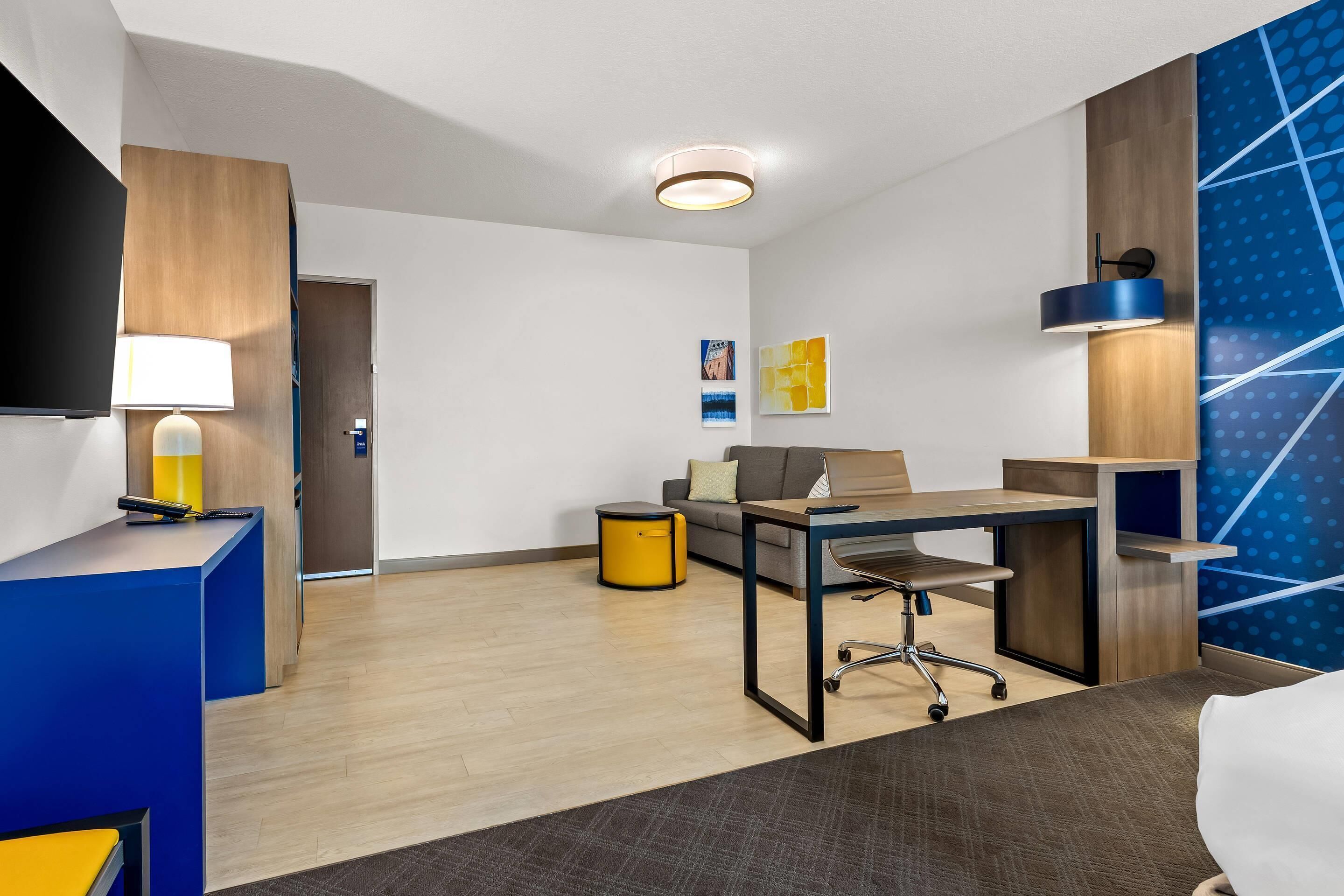 Una habitación de hotel moderna con escritorio, silla y cama. Cuenta con un mostrador de recepción con lámpara, una pequeña mesa redonda y un sofá. La habitación tiene piso de madera y una pared azul decorativa.