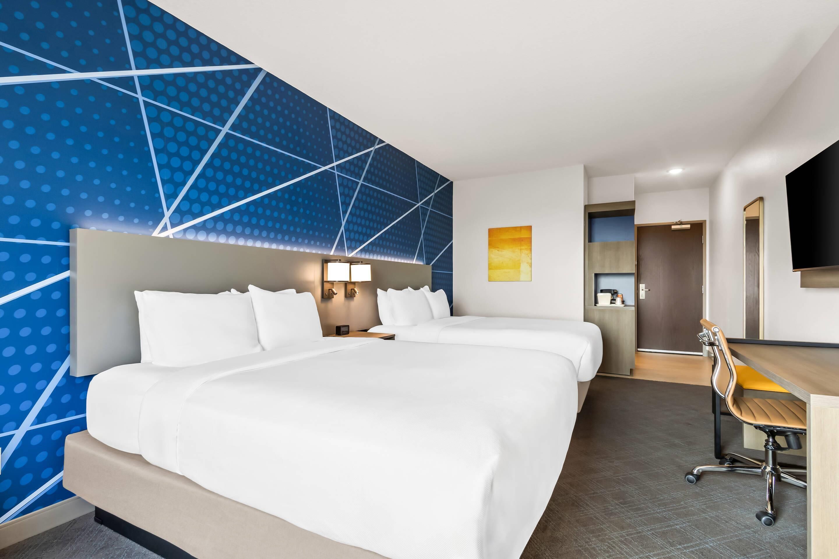 Habitación de hotel con una cama grande, un escritorio y una silla. Se observa una pared con un diseño geométrico y un cuadro.