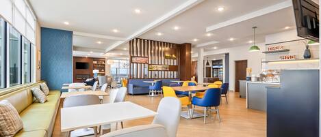 A modern dining area with a mix of seating options including tables and sofas, a bar area with a TV, and a kitchen visible in the background.