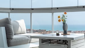 Ein modernes Zimmer mit einem weißen Sofa, einem kleinen Tisch mit Blumenvase und Blick auf Stadt und Meer durch große Fenster.