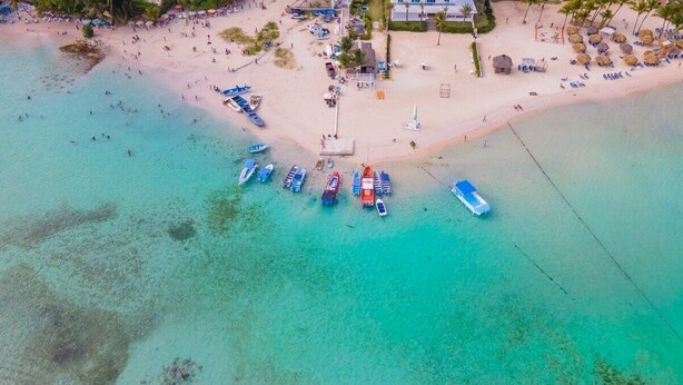 Ein Luftbild zeigt einen Strand mit mehreren angerten Booten, einen Sandstrand und einige Gebäude im Hintergrund.