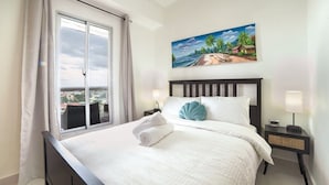 Ein ordentlich bezogenes Bett mit weißen Laken und einem blauen Kopfkissen, ein Strandbild an der Wand und ein Blick durch ein Fenster mit Vorhängen.