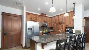 A kitchen with wooden cabinets, a stainless steel refrigerator, a microwave, a granite countertop, and bar stools. - Stylish, fully equipped 2-bed / 2-bath condo in Riverstone Village (Coeur d'Alene)