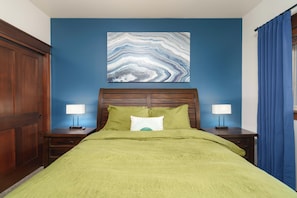 Ein Schlafzimmer mit einem grünen Bettbezug, zwei Nachttischen mit Lampen, einem Holzkopfende und einer blauen Akzentwand. Darüber hängt ein großes abstraktes Gemälde.