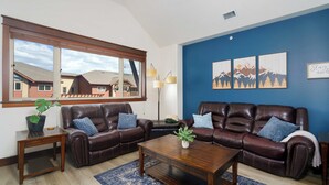 A living room with two leather sofas, a wooden coffee table, a floor lamp, a large window, and a blue accent wall with mountain artwork. - Stylish, fully equipped 2-bed / 2-bath condo in Riverstone Village (Coeur d'Alene)