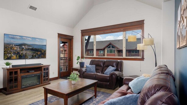 A living room with a fireplace, a large window, a brown leather sofa, a wooden coffee table, and a television. - Stylish, fully equipped 2-bed / 2-bath condo in Riverstone Village (Coeur d'Alene)