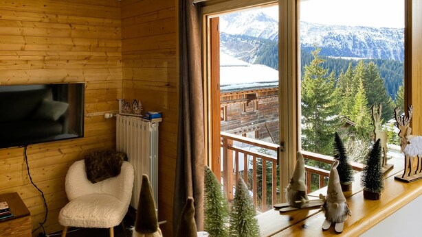 Una habitación acogedora con detalles de madera, un televisor montado, una silla blanca y vistas a montañas nevadas a través de amplias ventanas.
