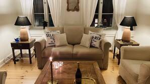 Una sala de estar acogedora con un sofá color beige, dos mesitas de noche con lámparas, una mesa de centro de madera con una vela, y almohadones decorativos.