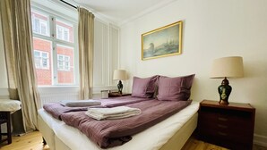 Una cama bien hecha con almohadas y una colcha, una mesita de noche con lámpara, una ventana con cortinas y un cuadro en la pared.