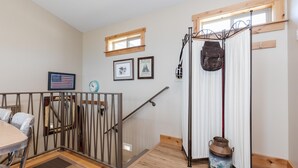 Una habitación con piso de madera, una mesa de comedor con sillas, una escalera con barandilla de metal, una ventana con marco de madera y una pared con cuadros enmarcados.