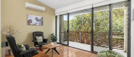 A spacious room with a wooden floor, a black leather chair, a wooden coffee table, and a green bean bag. There is a large sliding glass door leading to an outdoor balcony with a wooden fence and greenery.