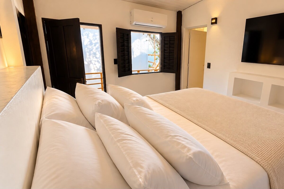 Una habitación de hotel con cama, almohadas, televisor y ventana con contraventanas.