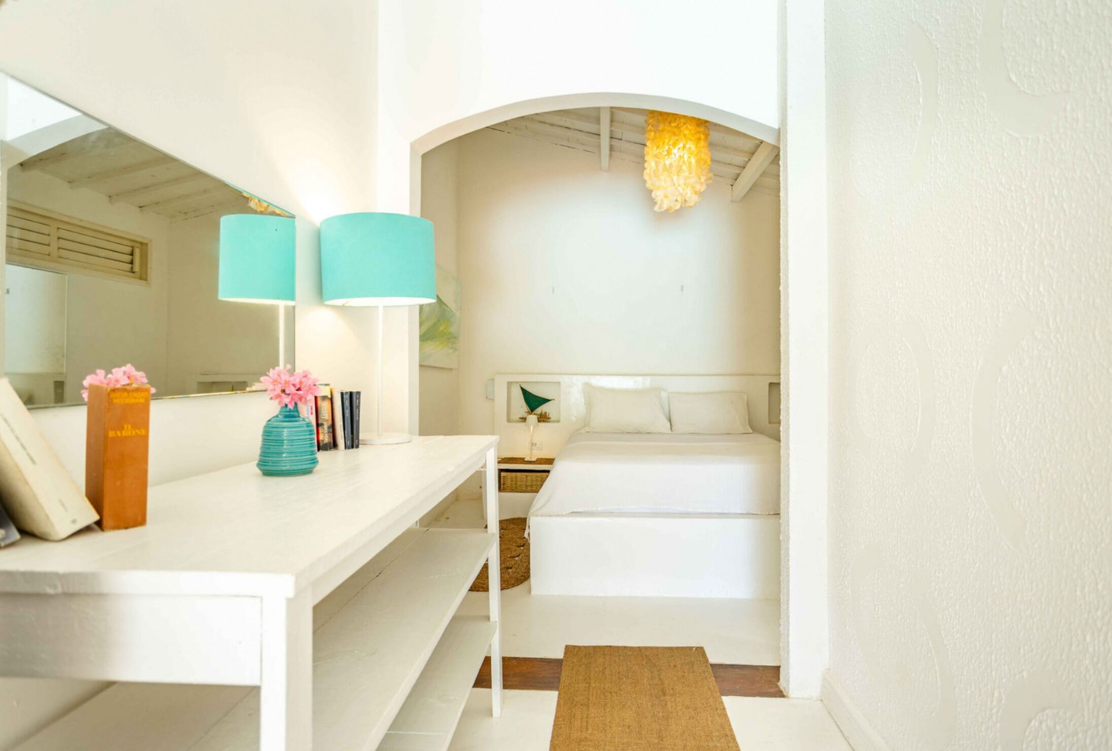 A bedroom with a white bed, a white desk with a lamp, and a mirror.