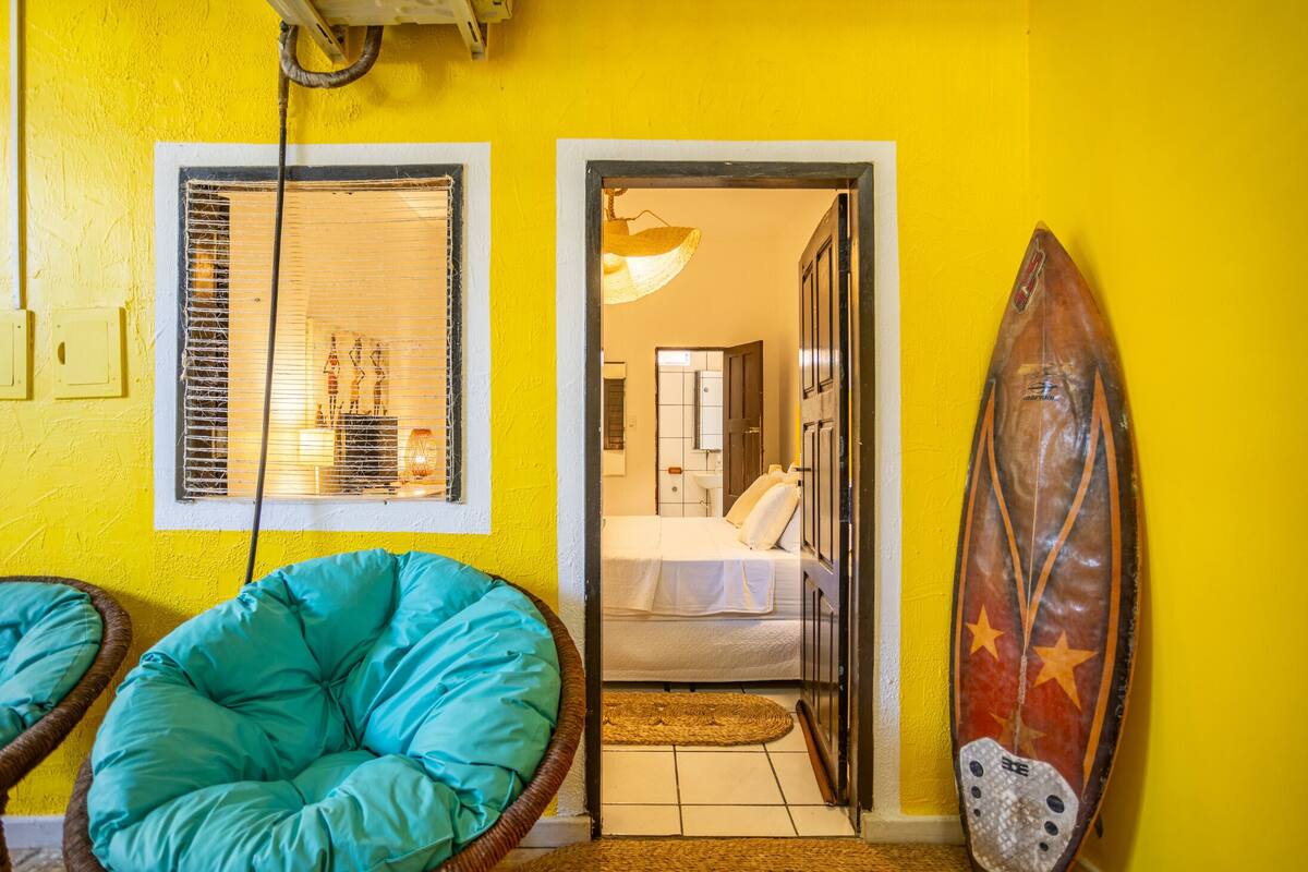 Ein Zimmer mit einem Bett, einem Sessel, einem Surfbrett und einem Fenster mit Jalousien.