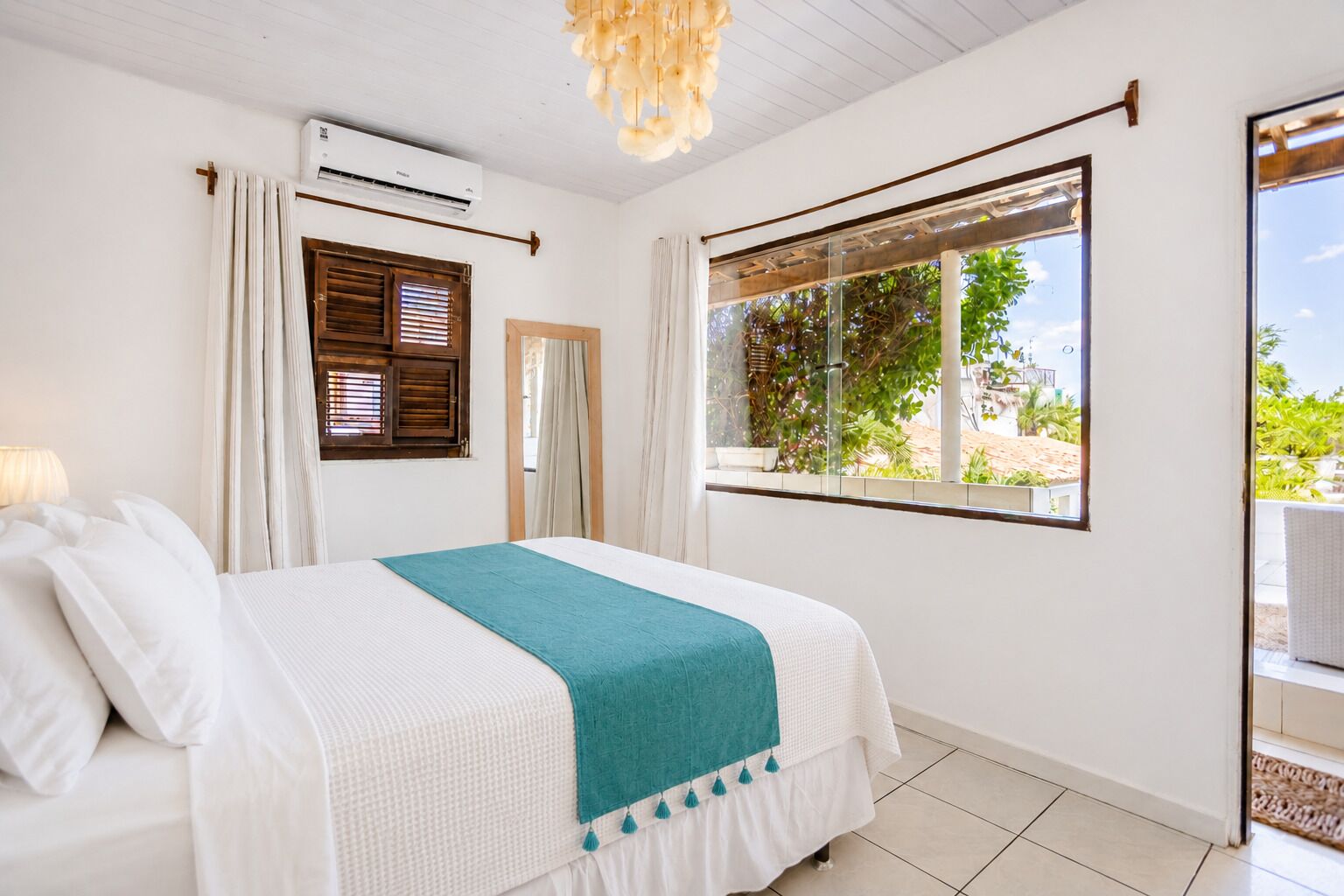 A bedroom with a bed, white curtains, a window with a view, and an air conditioning unit.