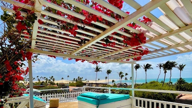 A covered outdoor seating area with wicker furniture and cushions, overlooking a beach and palm trees. - Pousada Duna Beach (Jijoca de Jericoacoara)