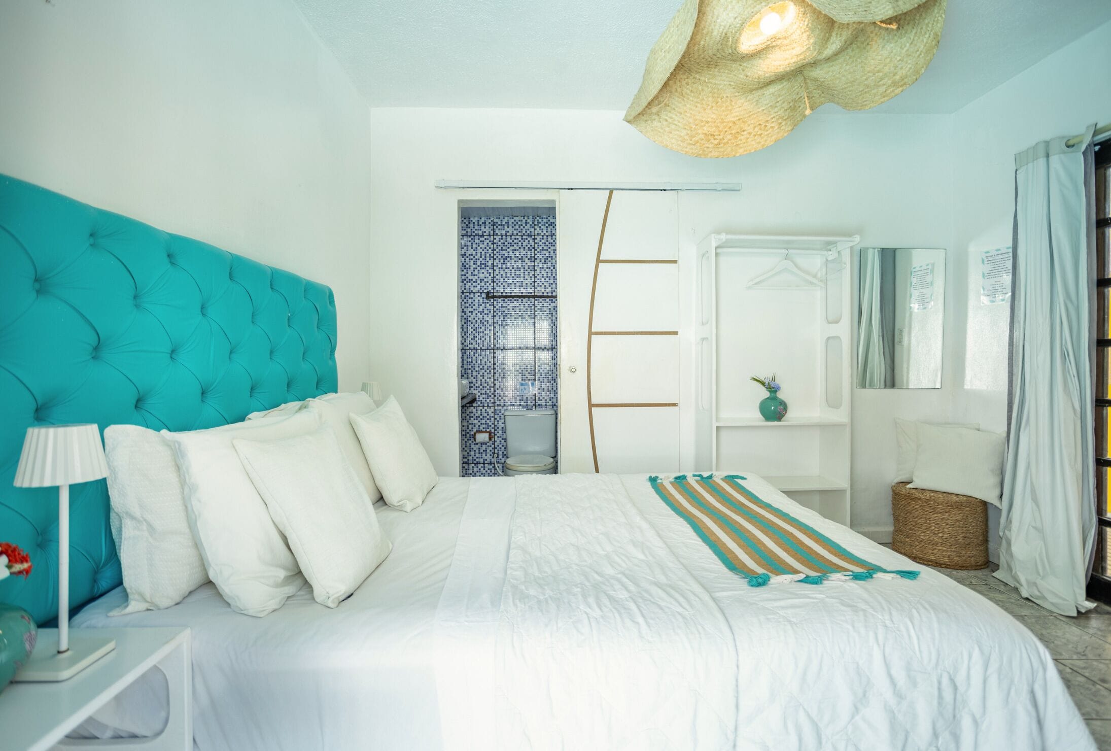 A bedroom with a teal headboard, white bedding, a striped throw blanket, a bedside lamp, a wicker basket, and a mirror on the wall.
