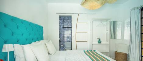 A bedroom with a teal headboard, white bedding, a striped throw blanket, a bedside lamp, a wicker basket, and a mirror on the wall.